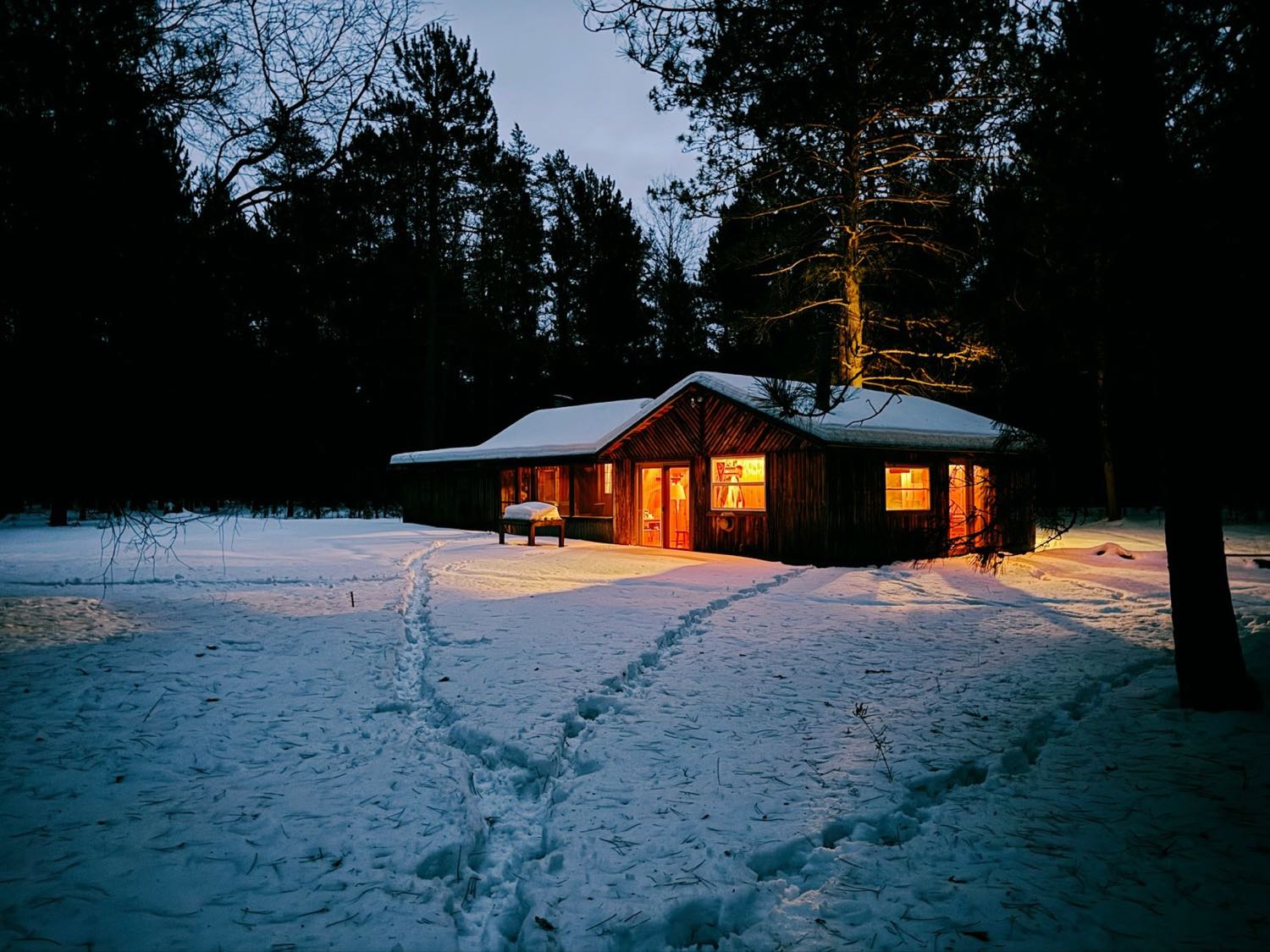 A low timber building with warmly lit windows standing in deep snow at dusk